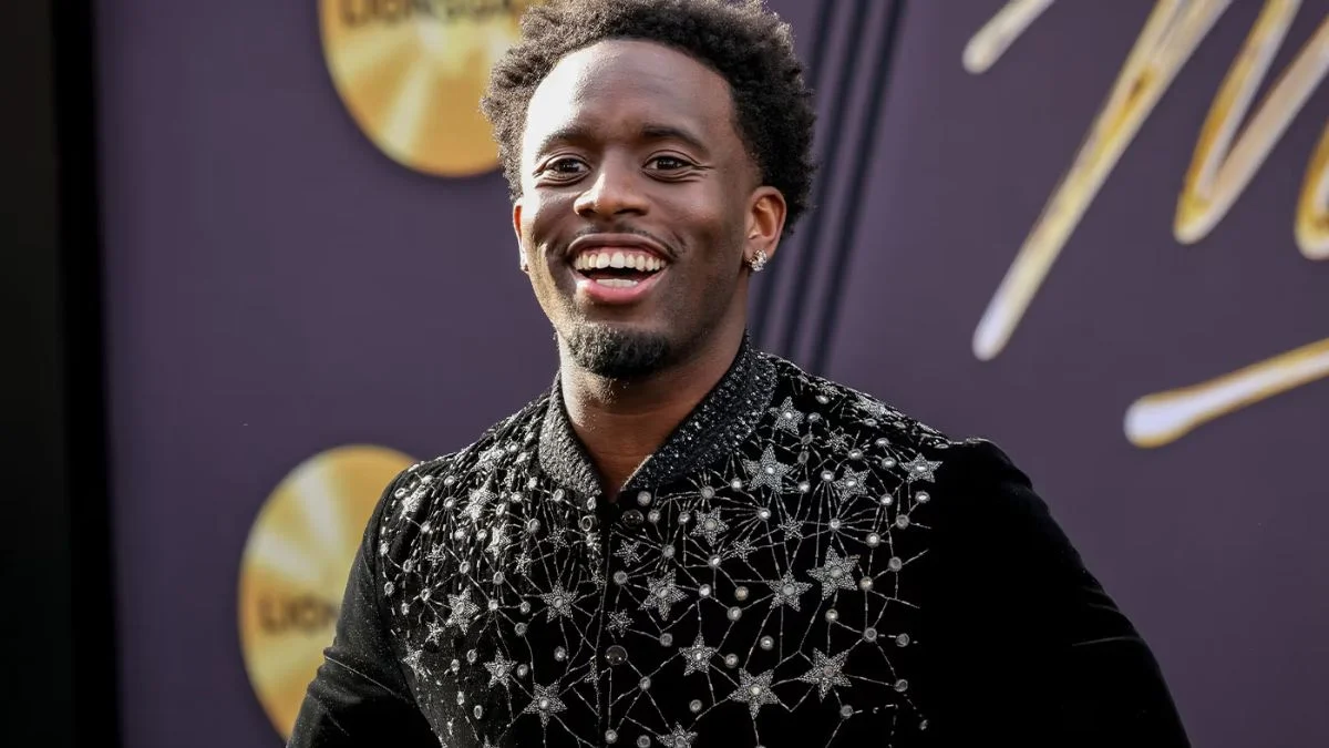 Close-up portrait of a smiling Kai Cenat wearing a black jacket with silver star embellishments on a red carpet.