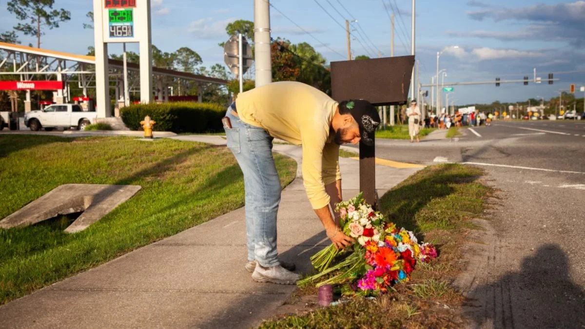 A man in a yellow shirt, blue jeans, and a black baseball cap bends down to place a large bouquet of colorful flowers at a roadside memorial sidewalk next to a busy road near a Wawa gas station.