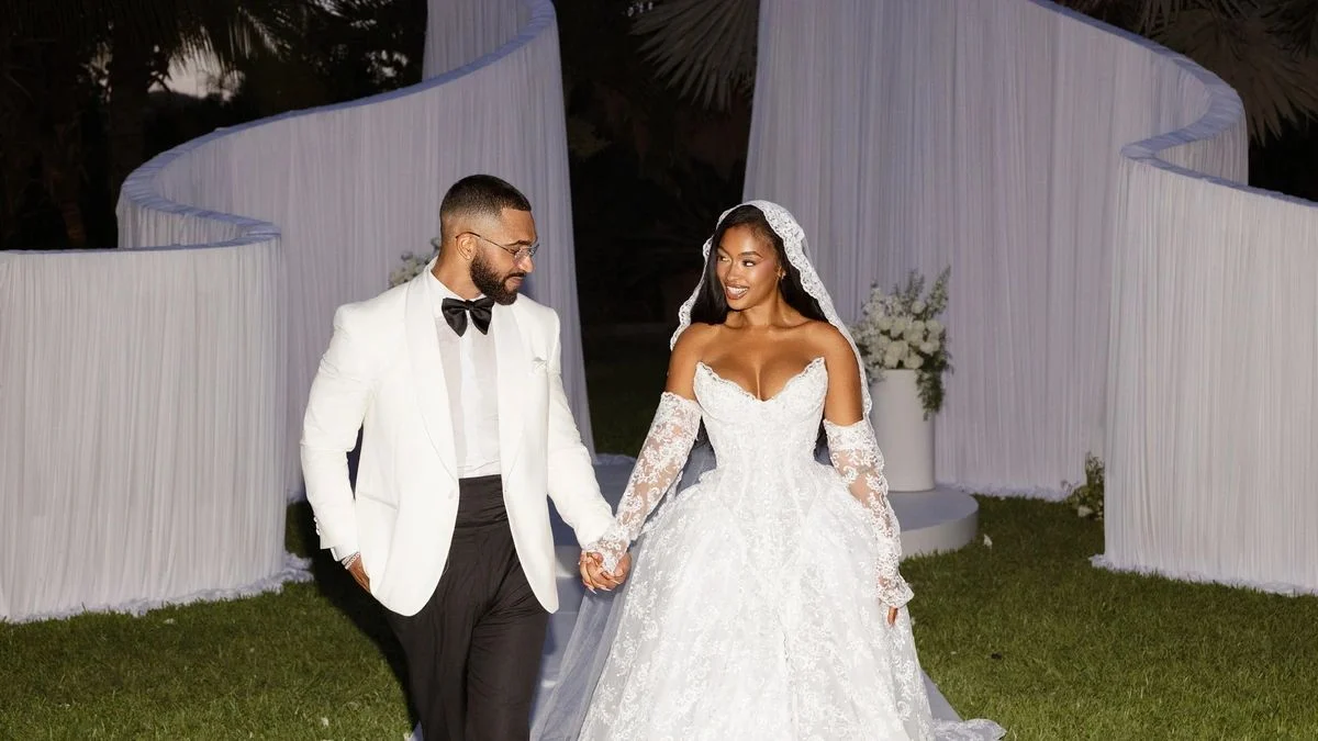 Newlyweds Tyler Lepley in a cream dinner jacket and black bow tie holding hands with Miracle Watts in an all-lace bridal gown and hooded veil at an outdoor resort venue.