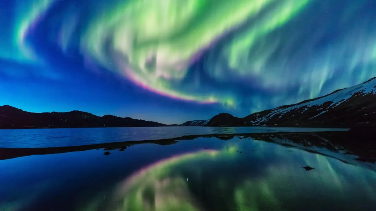 Bright green and purple northern lights stretching across the night sky and reflecting perfectly in a calm lake with snow-capped mountains in the background.