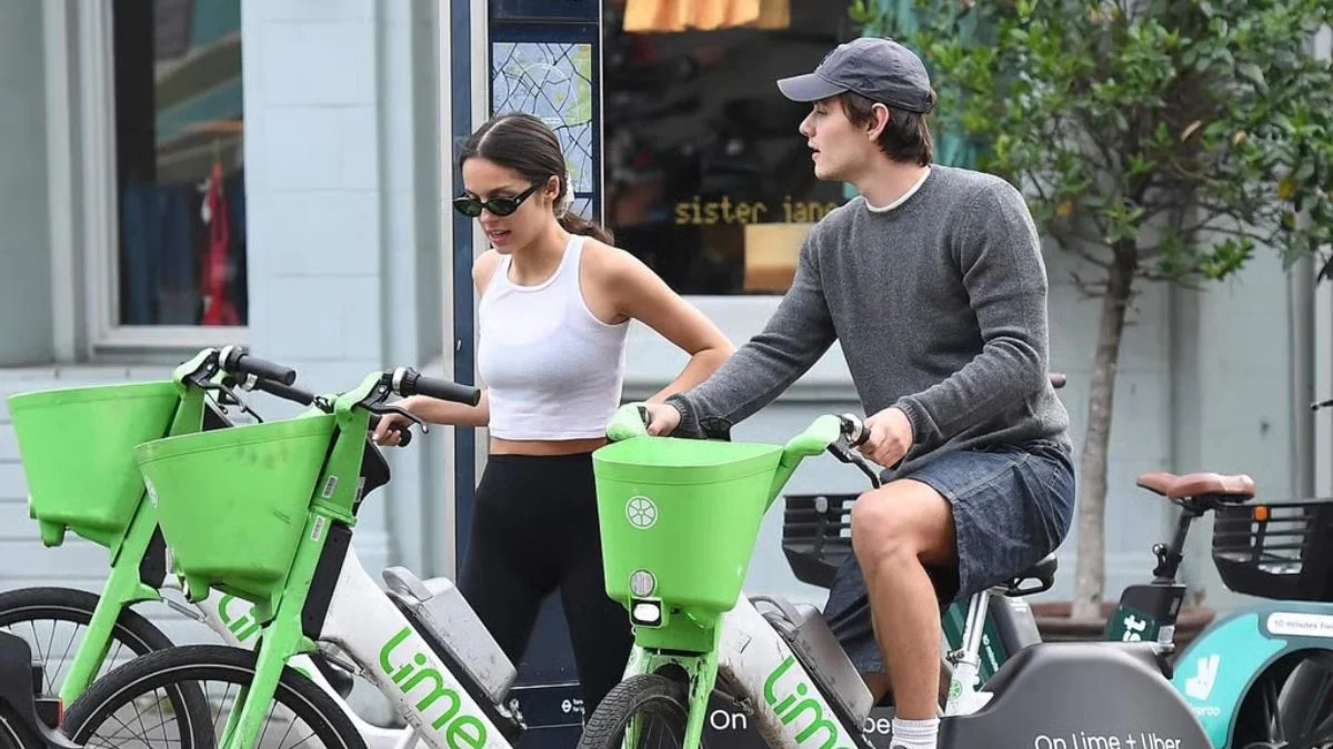 Olivia Rodrigo in a white crop top and sunglasses next to Louis Partridge as he sits on a green Lime electric bicycle on a city sidewalk.