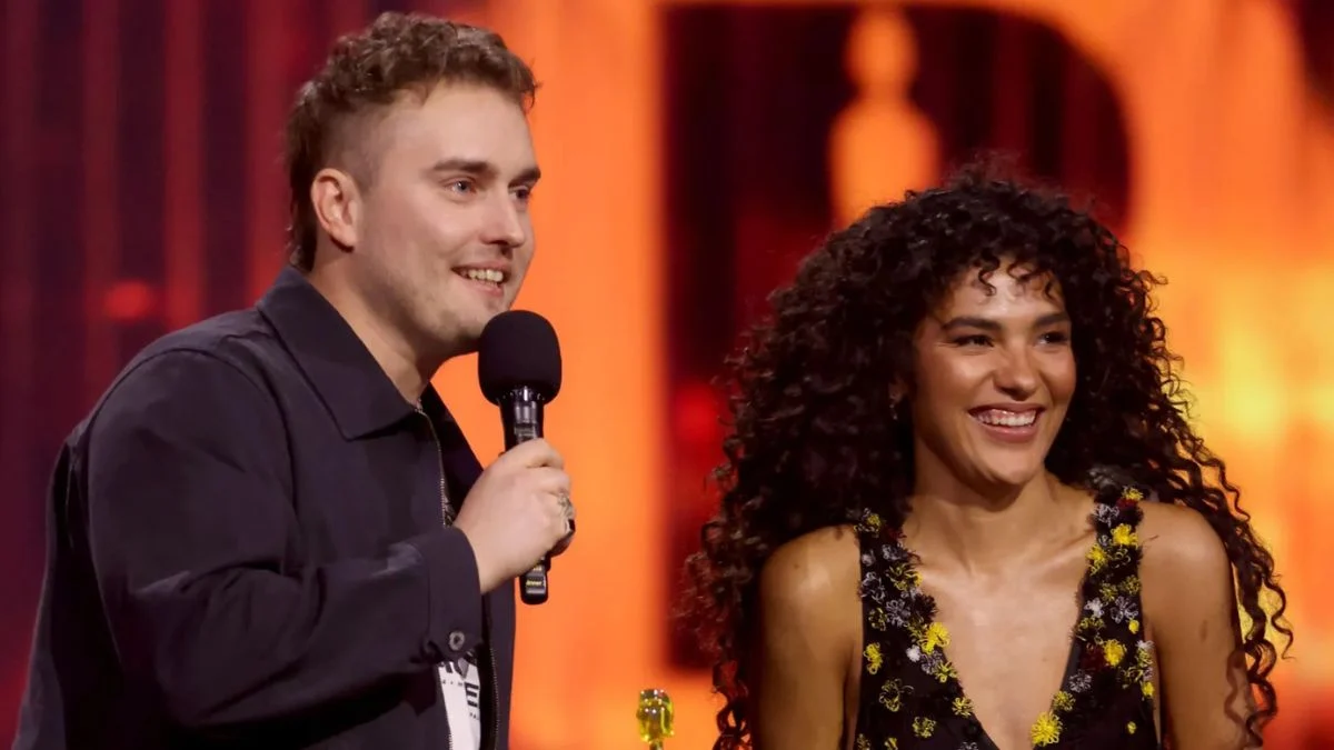 Sam Fender holding a microphone alongside a smiling Olivia Dean on stage at the Brit Awards against a warm orange background.