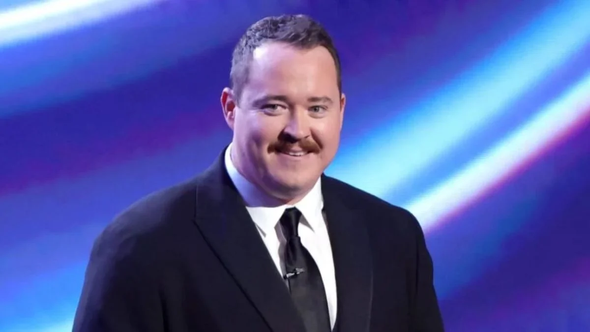 A close-up shot of comedian Shane Gillis smiling on stage in a dark suit jacket and tie against a vibrant blue and purple abstract lighting background.