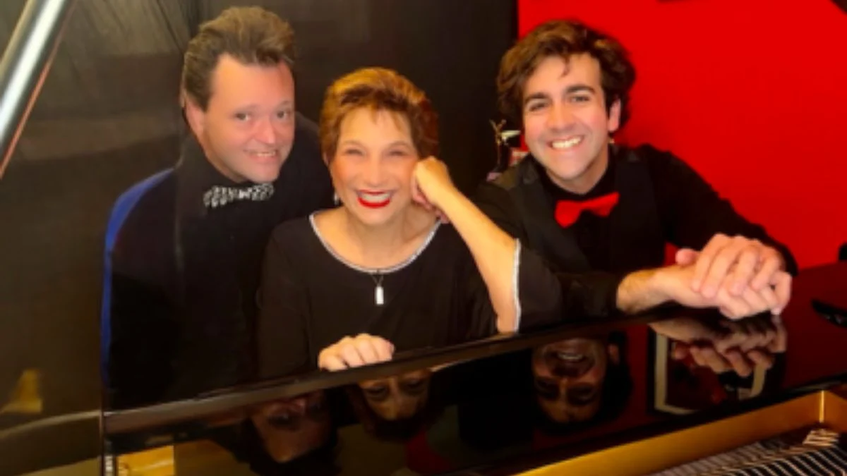 Pianists Julian Bond, Carol J. Stein, and Michael Mastroianni smiling together while leaning over the shiny glossy lid of a black grand piano.
