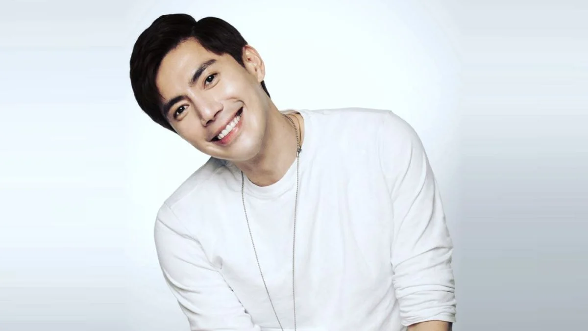 A studio portrait of Taiwanese actor Wang Kai smiling and tilting his head while wearing a long-sleeved white shirt and a thin silver chain necklace against a light grey gradient background.