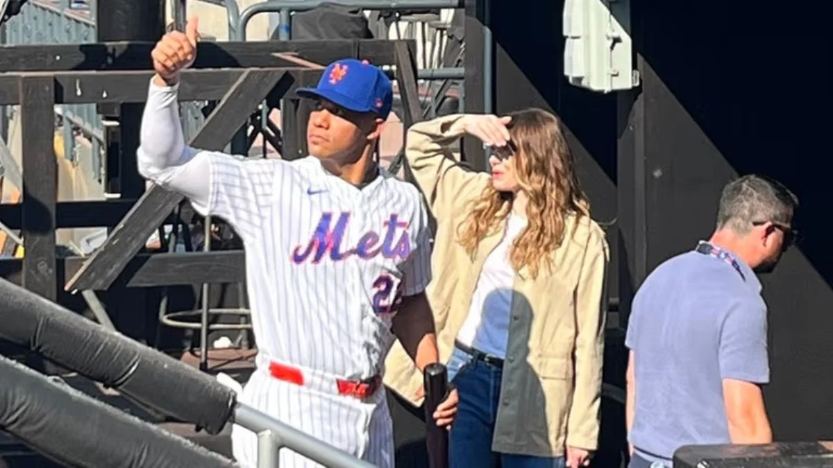 New York Mets player Juan Soto in a white pinstripe uniform giving a thumbs up next to actress Emma Stone in a tan jacket shielding her eyes from the sun in the dugout at Citi Field.