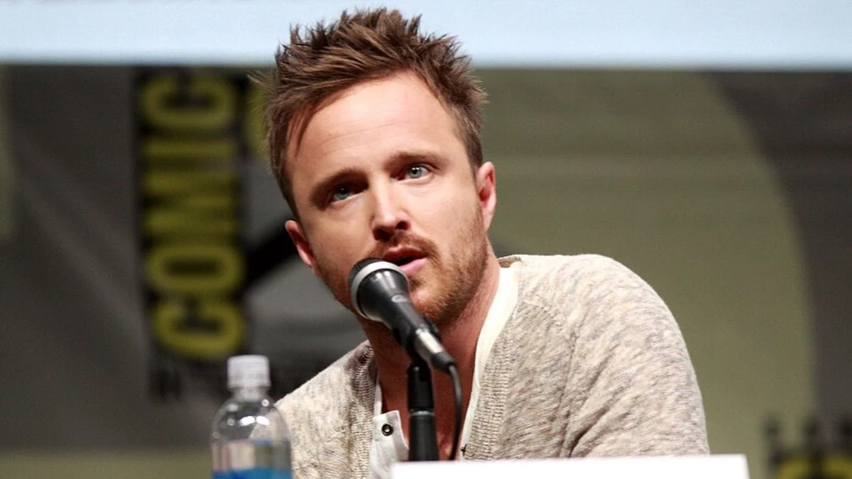 Aaron Paul speaking into a microphone on stage at a convention panel
