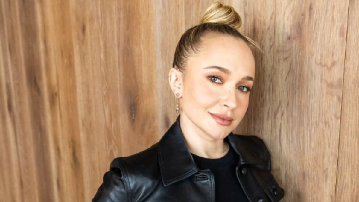 Hayden Panettiere with hair styled in a top bun leaning against a wooden wall in a black leather jacket.