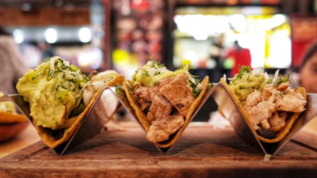 Trio of freshly made Tex-Mex tacos loaded with grilled meat, guacamole, and herbs on a metal taco holder.
