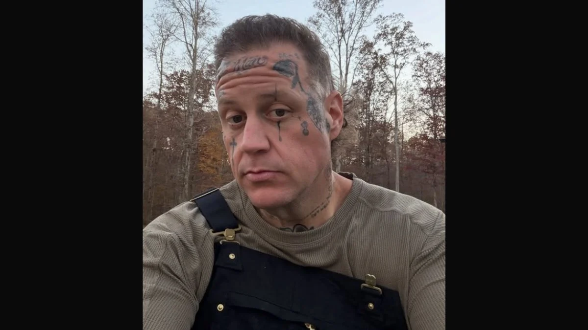 Country-rap star Jelly Roll taking a selfie outdoors wearing dark overalls and a tan waffle-knit shirt.
