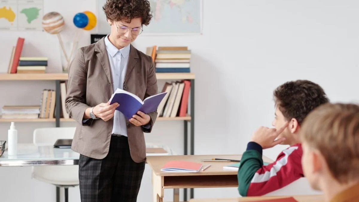 Teacher reading from a notebook while standing in front of students seated at classroom desks