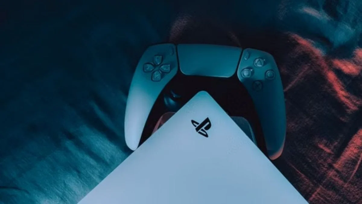 A PlayStation 5 console edge and a DualSense controller sitting on a dark surface lit with dramatic blue and red ambient lighting.