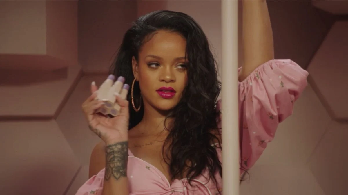 Rihanna posing in a pink off-the-shoulder outfit with bold magenta lip color and hoop earrings.