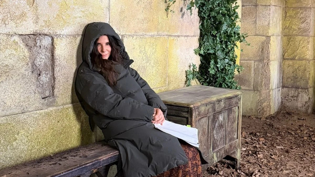 Sandra Bullock leaning against a stone wall while wearing a long hooded puffer coat and holding scripts on set.
