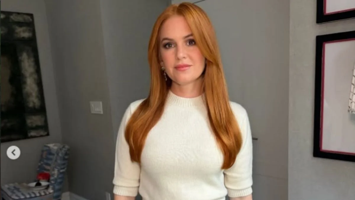 Actress Isla Fisher with red hair wearing a white fitted sweater indoors looking at the camera.