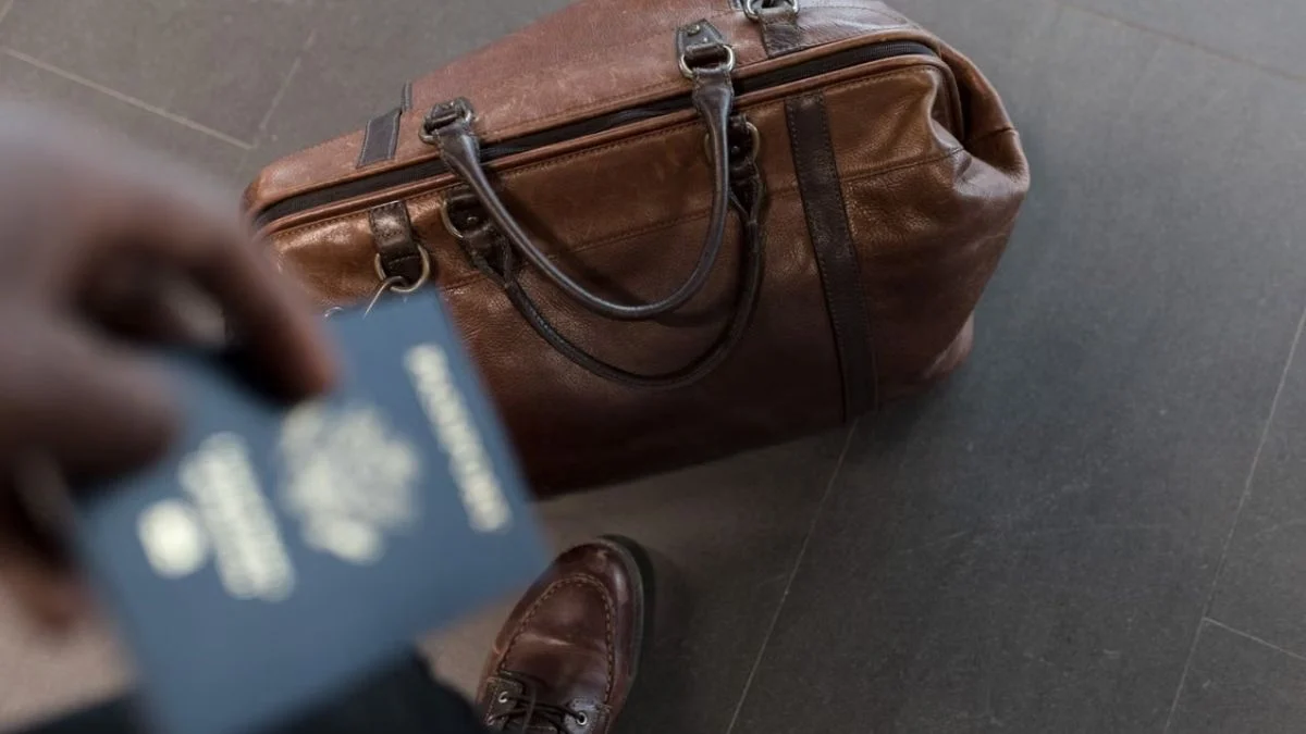 First-person view of a traveller holding a passport in front of a brown leather carry-on bag resting on the floor.
