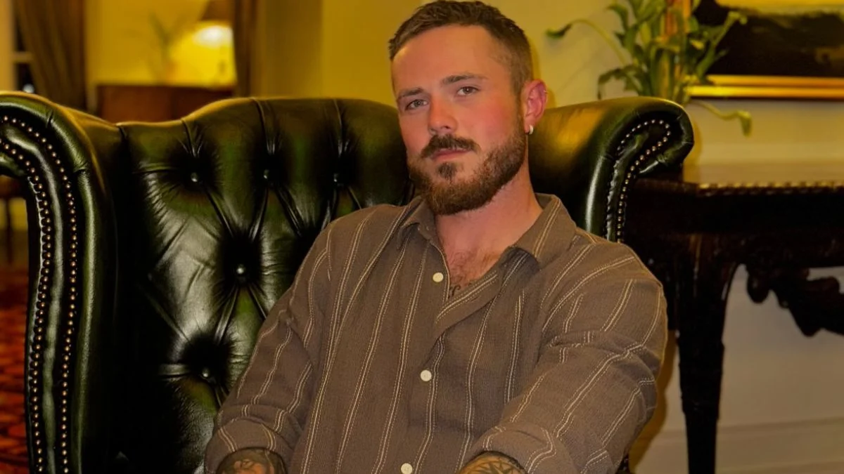 Below Deck Mediterranean star Nathan Gallagher posing in a striped button-down shirt while sitting in a dark leather armchair.