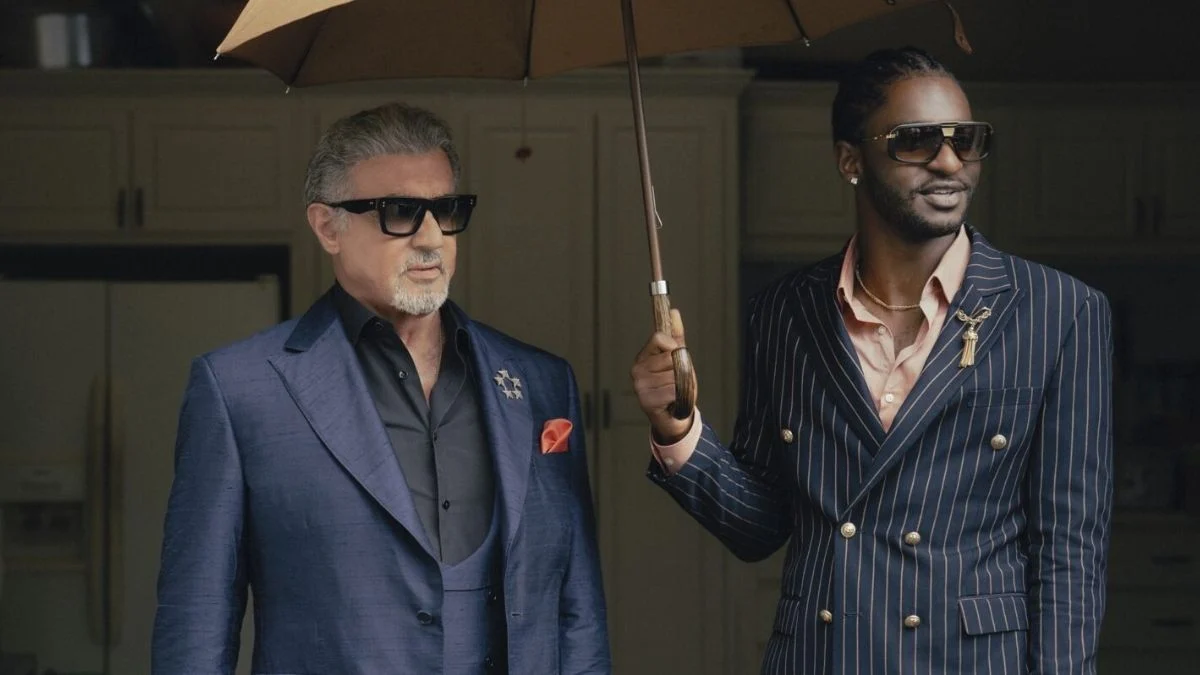 Sylvester Stallone wearing sunglasses standing next to a co-star holding an umbrella on set