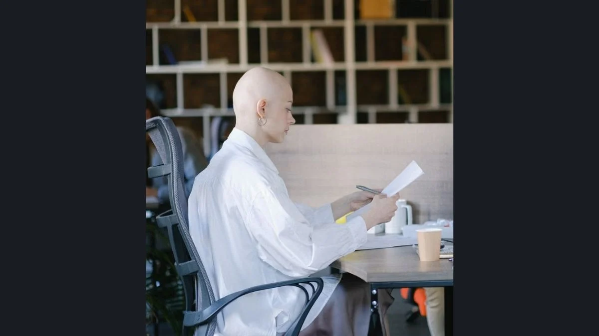 Profile view of a bald woman sitting in a white top at a desk, carefully reading a document.