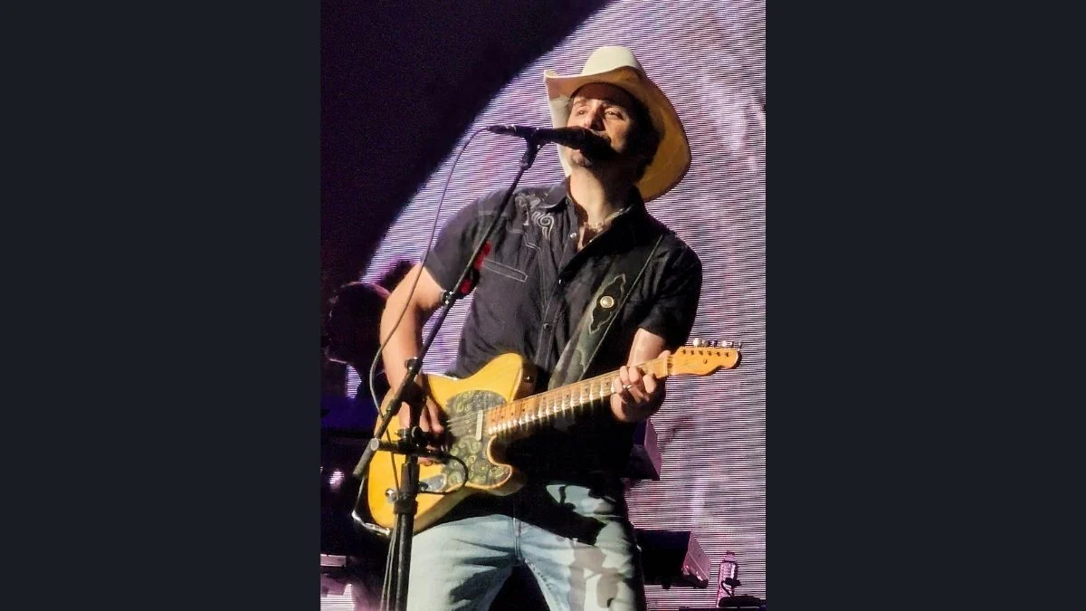 Brad Paisley wearing a white cowboy hat and black shirt while playing an electric guitar and singing into a microphone on stage.