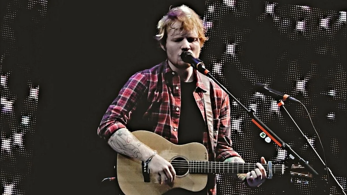 Ed Sheeran singing into a microphone while playing an acoustic guitar on stage in a red plaid shirt.