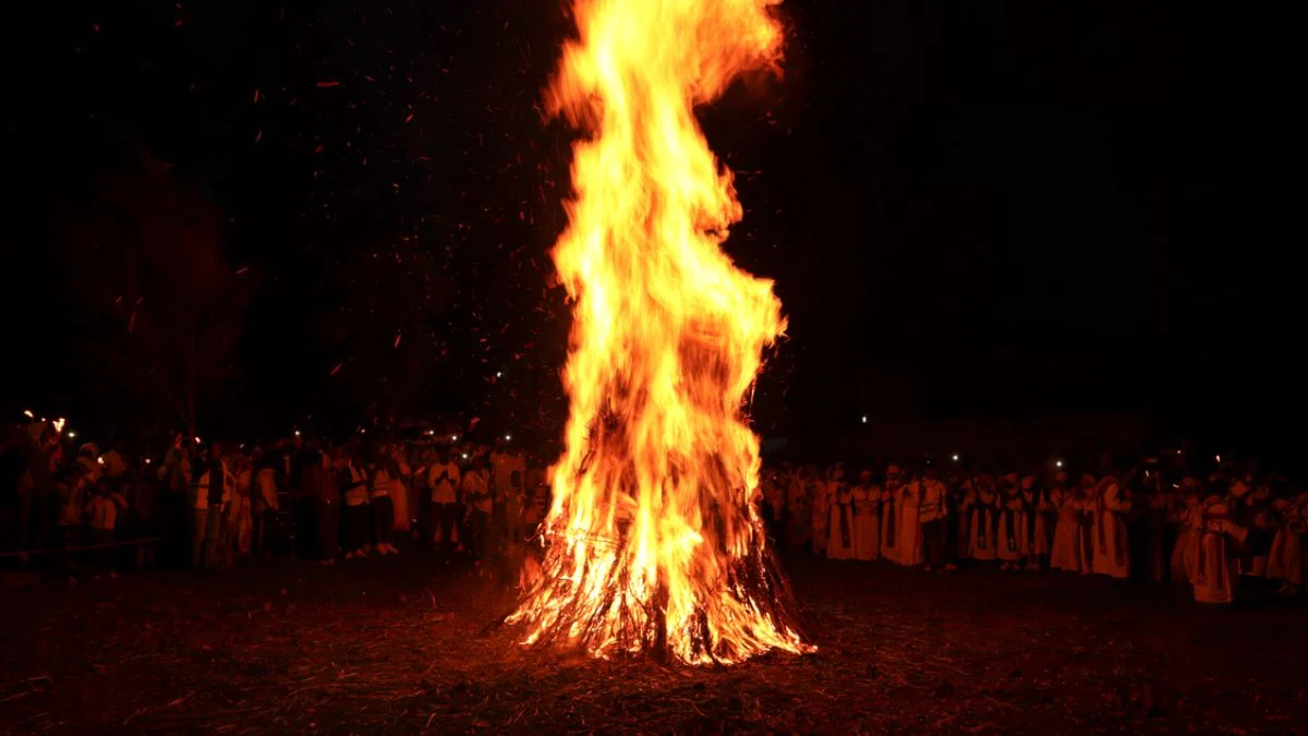 Large bonfire with high orange flames burning brightly in the dark at night surrounded by crowd silhouettes.