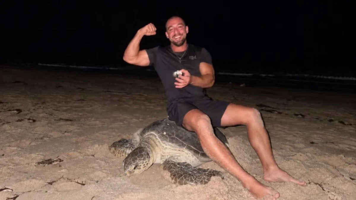 Vadym Oleynik smiling and flexing his bicep while sitting on the shell of an adult green sea turtle on a beach at night.