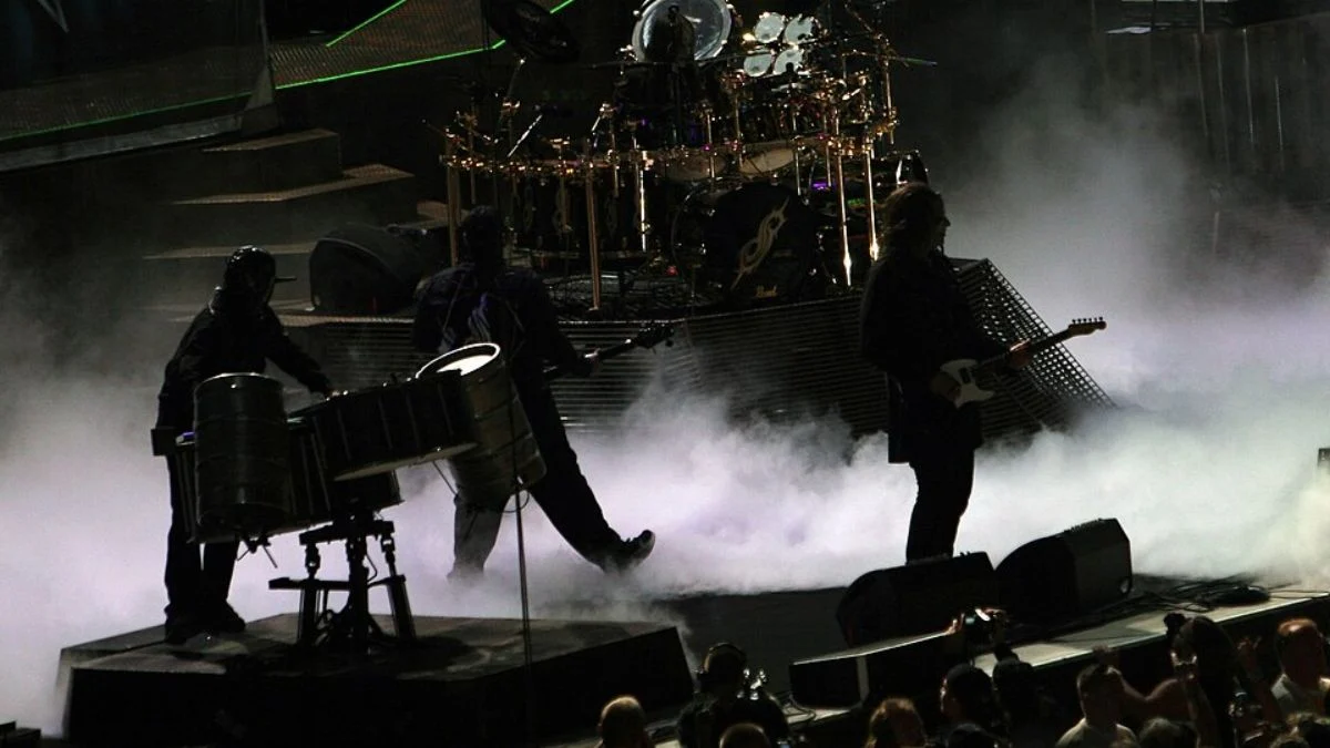 Members of Slipknot performing live on stage surrounded by heavy fog and dramatic stage lighting during a concert.