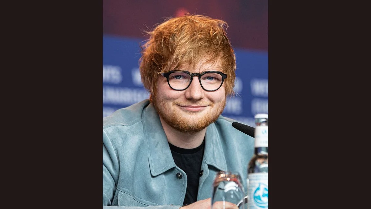 Ed Sheeran wearing black-rimmed glasses and a light blue jacket smiling during a press conference.