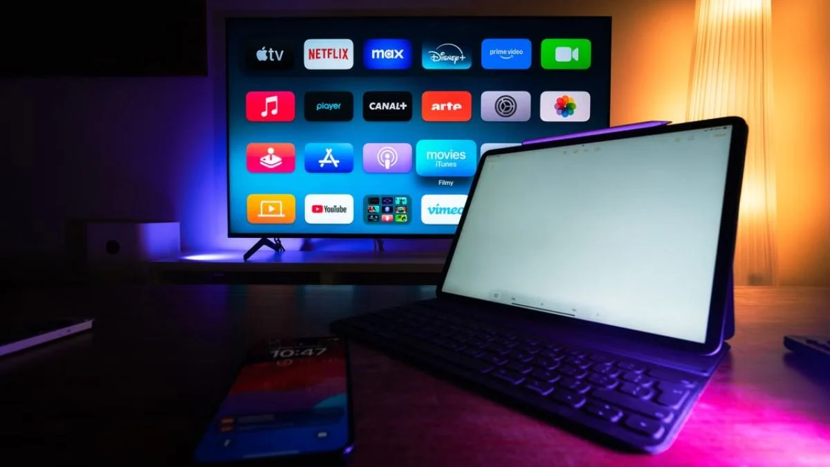 A tablet and smartphone on a desk in front of a smart TV displaying media apps including YouTube