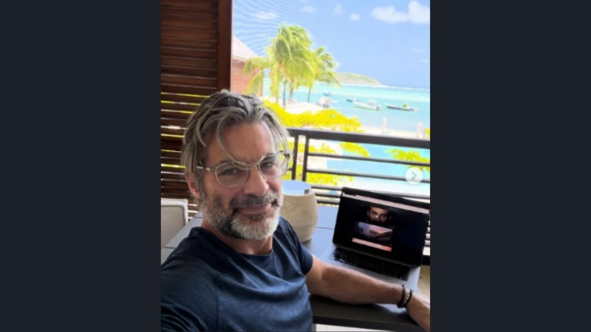 Jim Curtis wearing glasses and a navy blue t-shirt taking a selfie outdoors with an open laptop next to him overlooking a tropical beach and ocean.