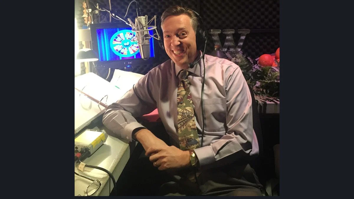 Wheel of Fortune announcer Jim Thornton sitting at a recording booth microphone smiling.