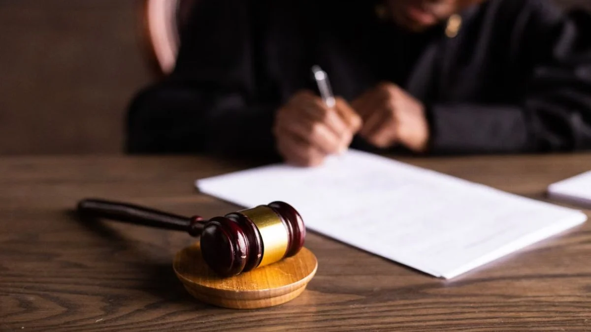 Wooden judge's gavel and sound block sitting on a desk as a judge writes on court documents during a legal hearing.