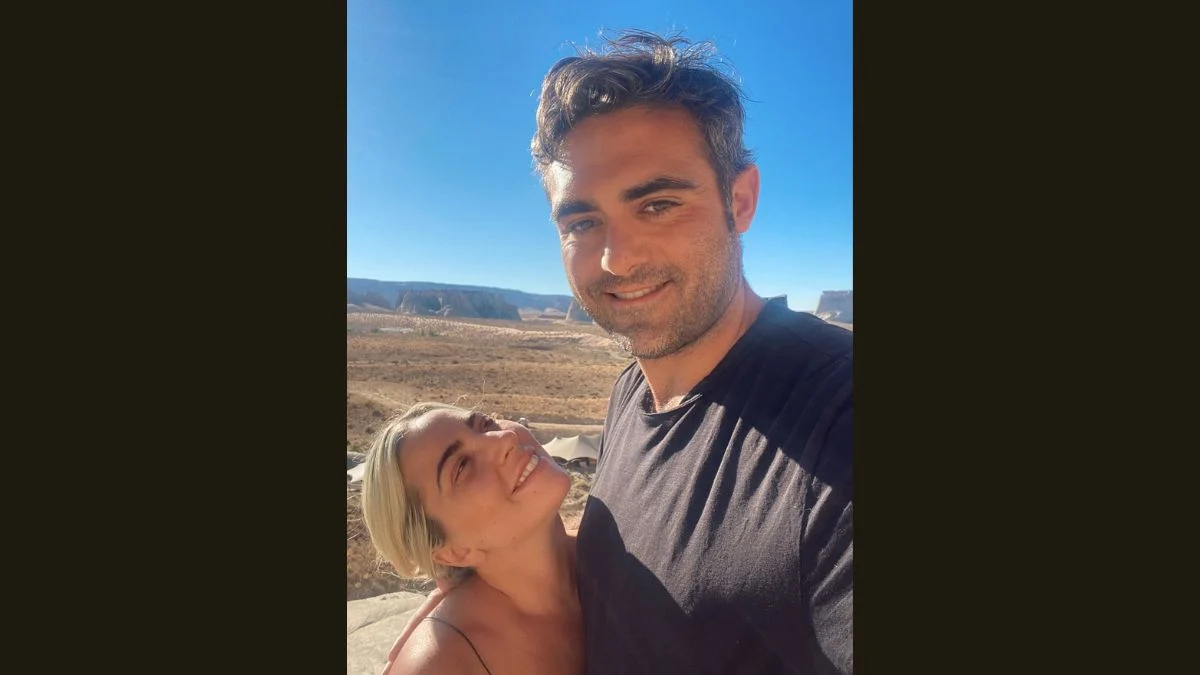 Lady Gaga smiling softly next to her fiancé Michael Polansky under a clear blue sky in a desert landscape.