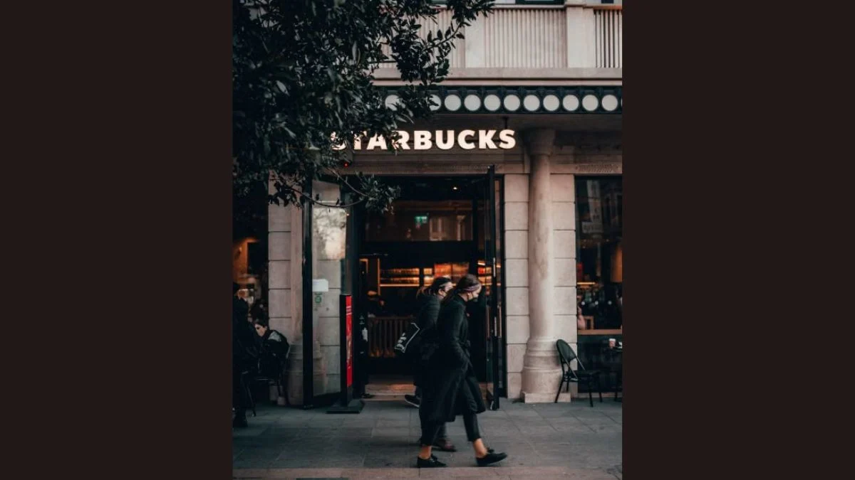 Pedestrians walking past an illuminated Starbucks storefront during the evening.