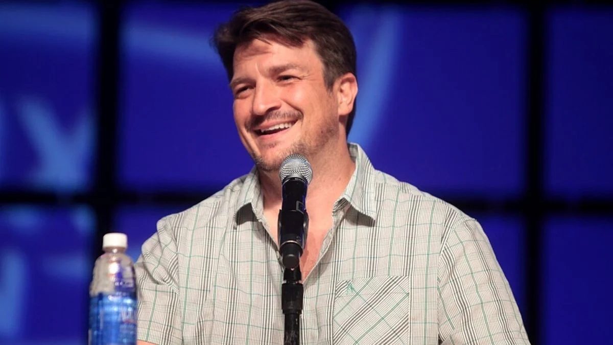 Actor Nathan Fillion smiling while speaking into a microphone on stage at a convention panel event, wearing a short-sleeved plaid shirt.