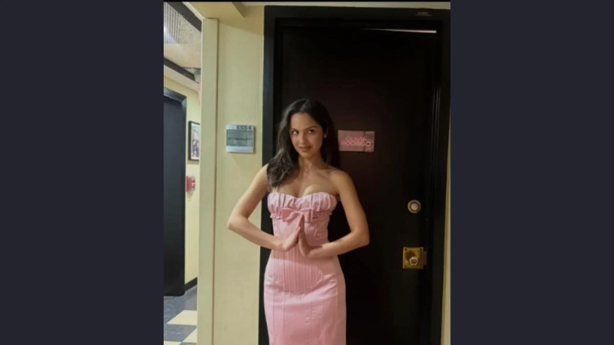 Olivia Rodrigo posing backstage in a strapless pink dress in front of her dressing room door.