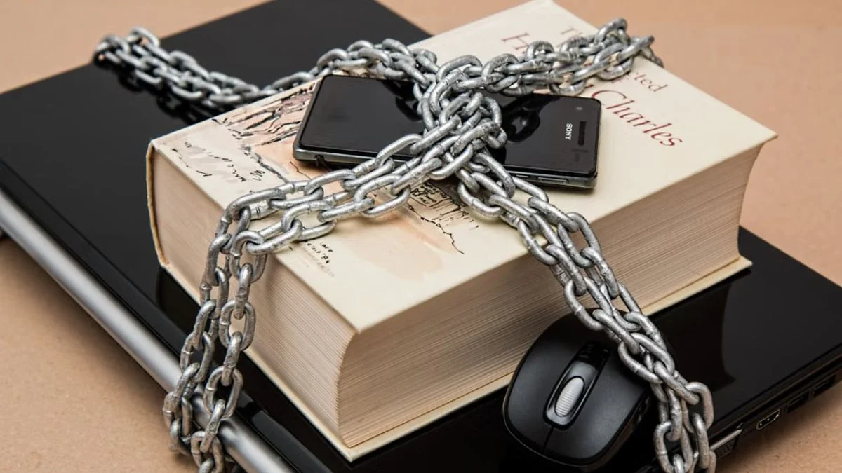 A heavy metal chain wrapped around a book, a smartphone, and a computer mouse on top of a laptop, representing digital lock, copyright, and AI publishing restrictions.