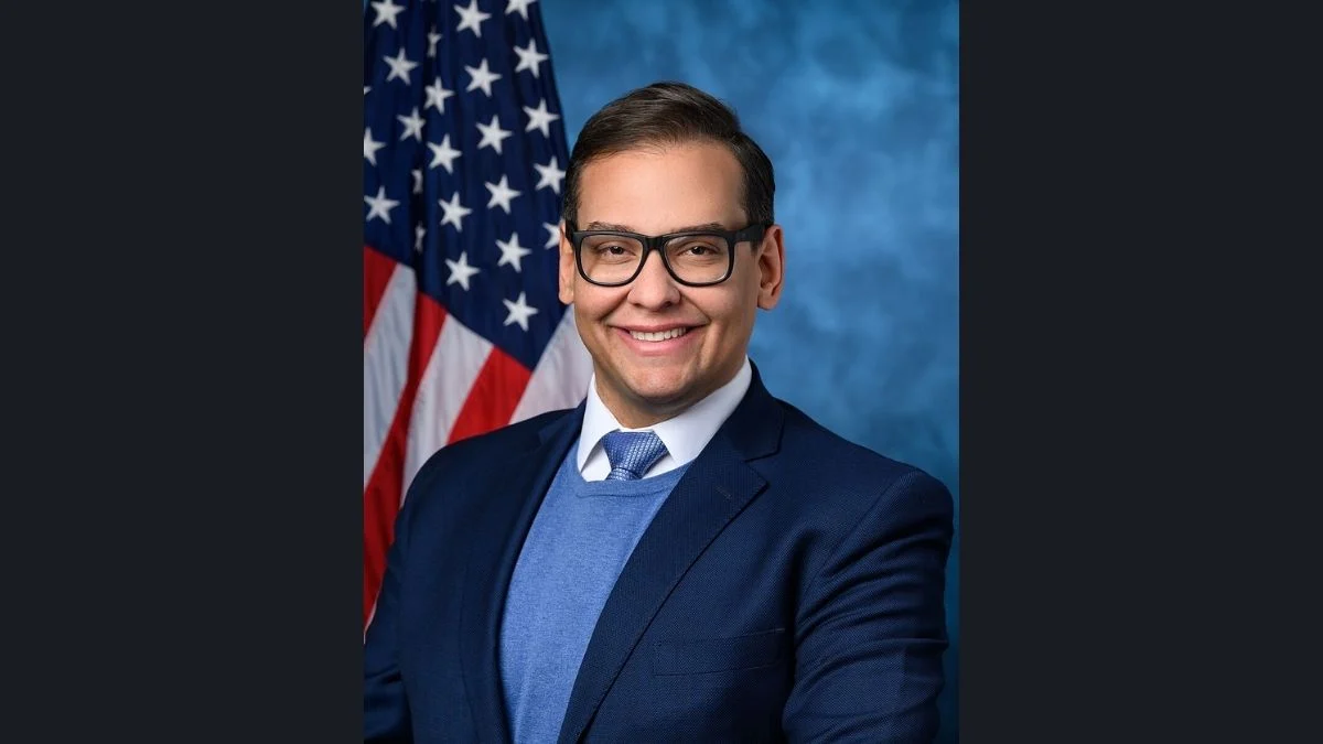 Official portrait of former U.S. Representative George Santos smiling while wearing black-framed glasses and a suit in front of an American flag.