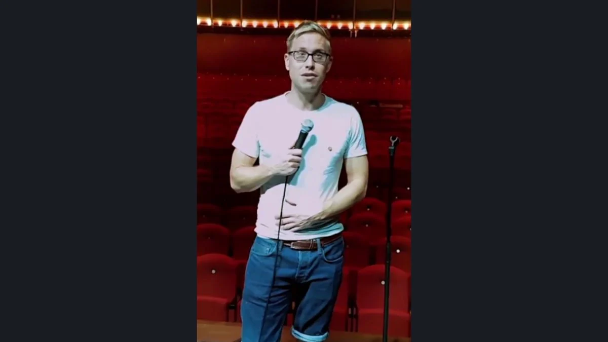 Comedian Russell Howard holding a microphone on stage in front of red theatre seating.