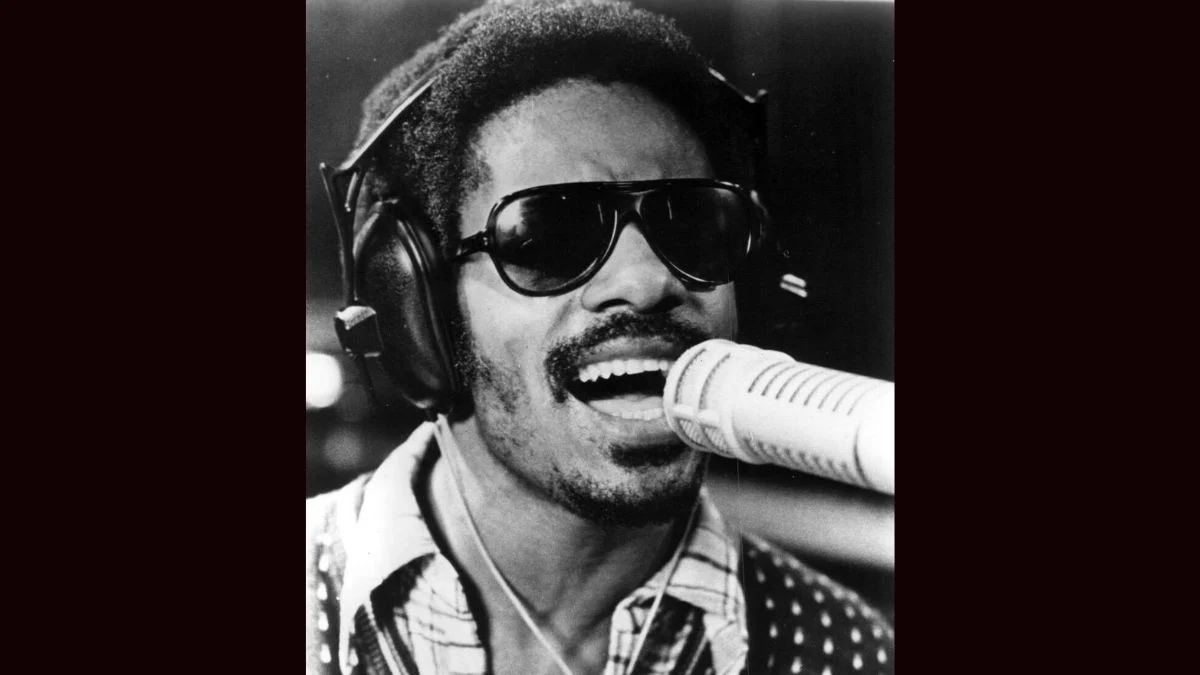 Classic black-and-white portrait of Stevie Wonder wearing sunglasses and headphones singing into a studio microphone.