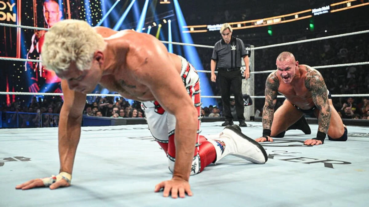 Cody Rhodes crawling on hands and knees in the foreground while Randy Orton kneels behind him shouting in the WWE ring.