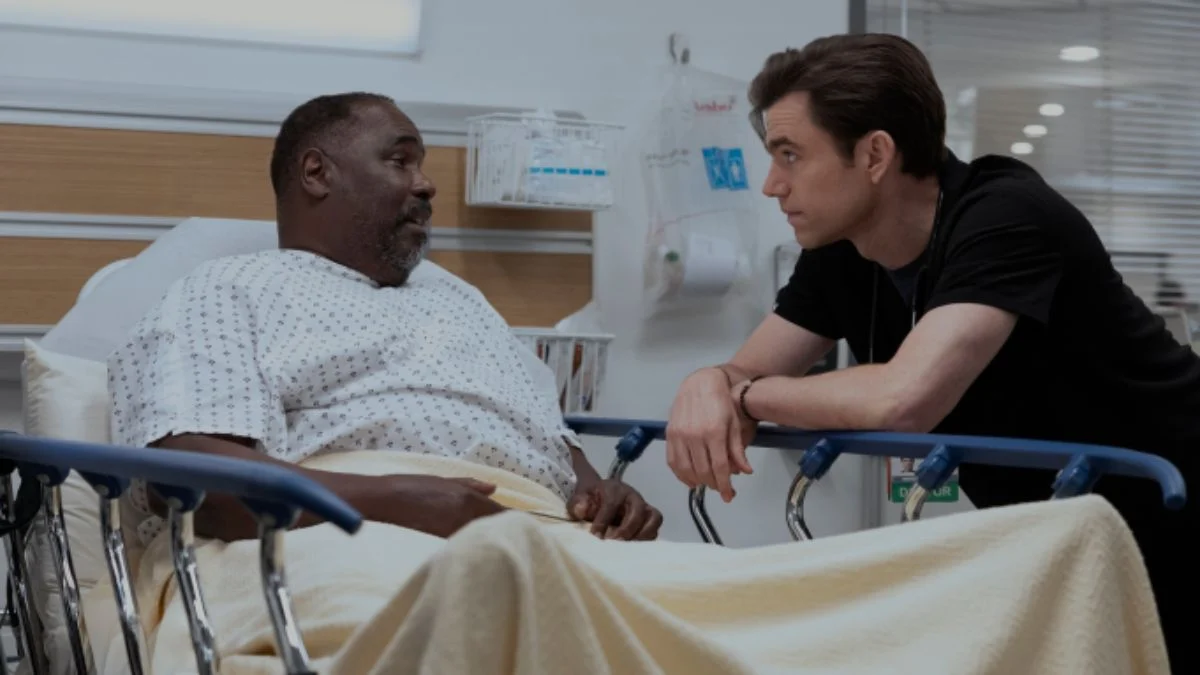 Actor Ernest Harden Jr sitting in a hospital gown on a gurney speaking to a male doctor leaning in to listen in a scene from The Pitt.