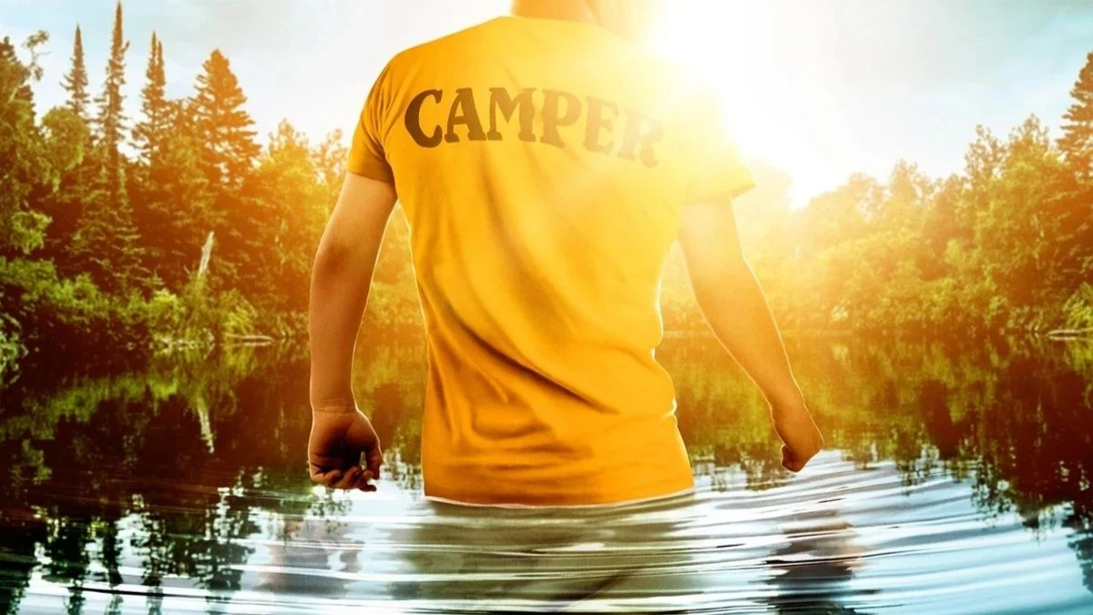 Rear view of a young camper wearing a bright yellow shirt with CAMPER printed on the back, standing knee-deep in water at sunny Crystal Lake.