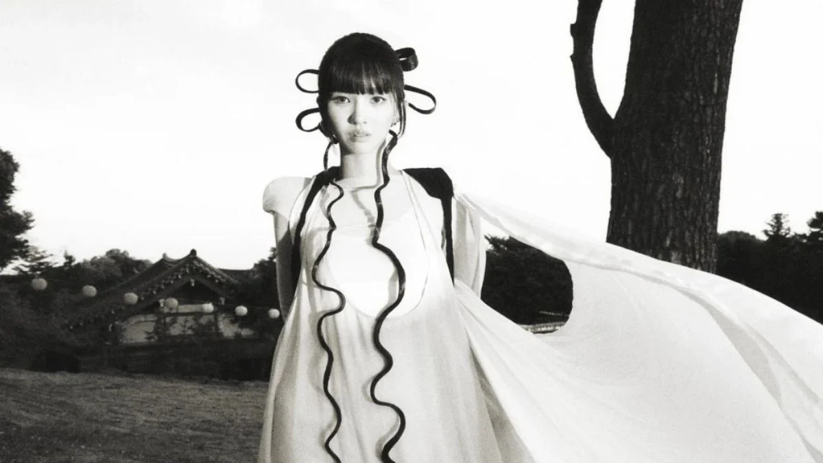 Black and white promotional concept photo of i-dle's Soyeon in a flowing white dress with dramatic coiled pigtails, standing outdoors near a tree and traditional building.