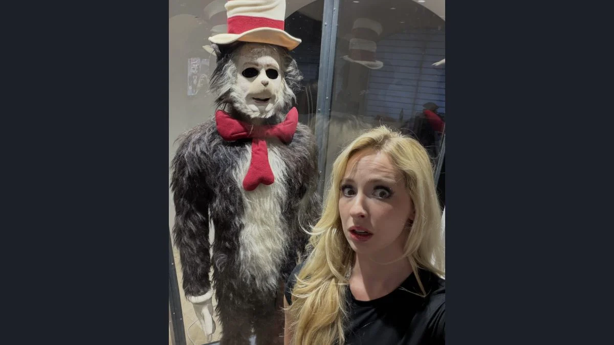 Natalie Reynolds posing next to the screen-used 2003 Cat in the Hat costume inside a glass display case.