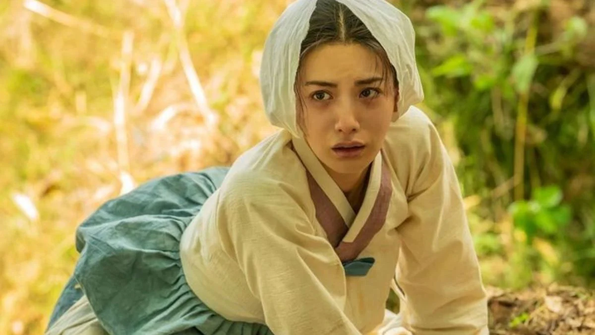 Nana as Hee Yeon in the Joseon-era Netflix drama The Scandal, wearing traditional hanbok and a head covering while kneeling outdoors.