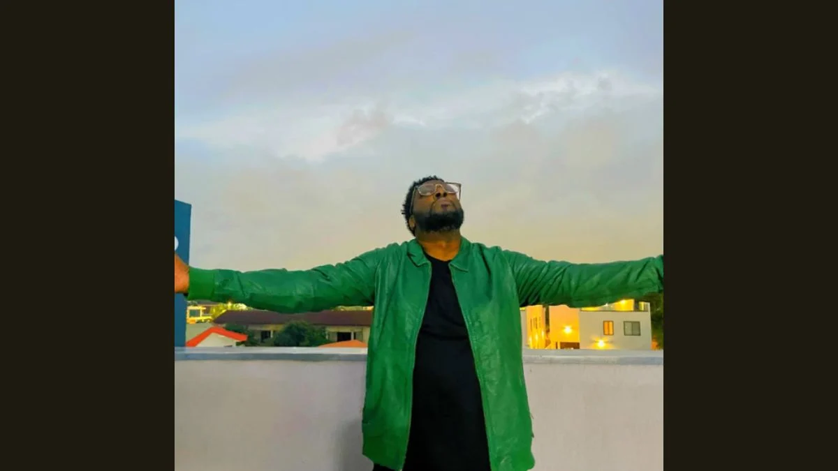British rap pioneer Sway DaSafo wearing glasses and a green jacket with arms outstretched under an open sky.