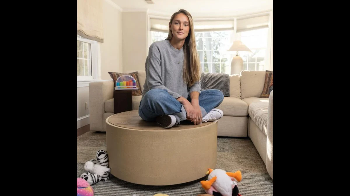 Kylie Kelce sitting cross-legged on an Ottoman coffee table in her living room surrounded by children's toys