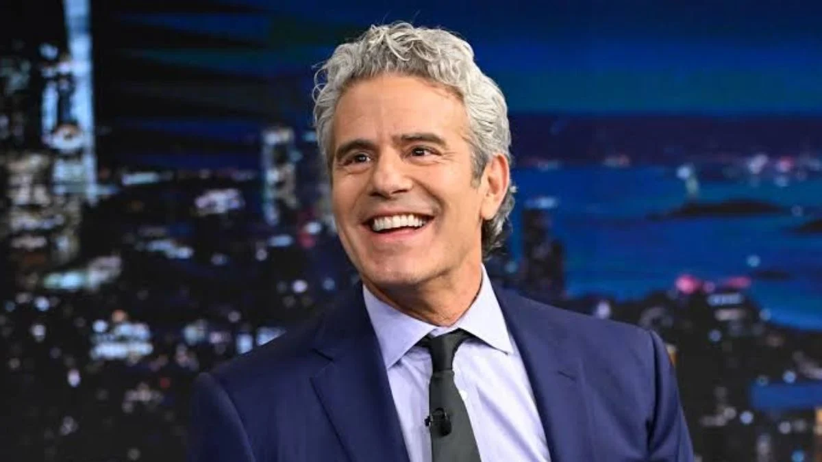 Andy Cohen smiling on a late-night talk show set wearing a navy suit and black tie